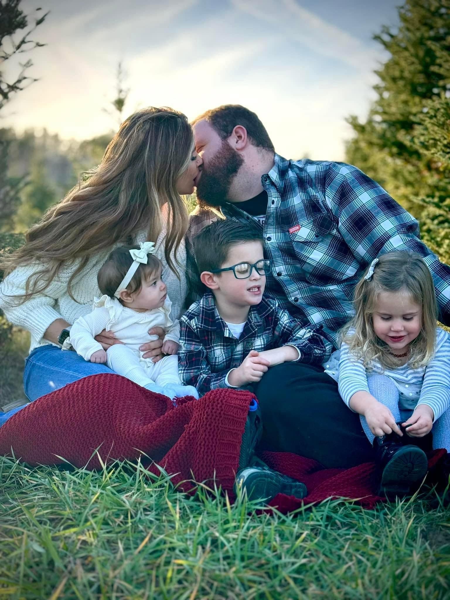 Parents kiss while sitting on a red blanket outdoors with their three young children.