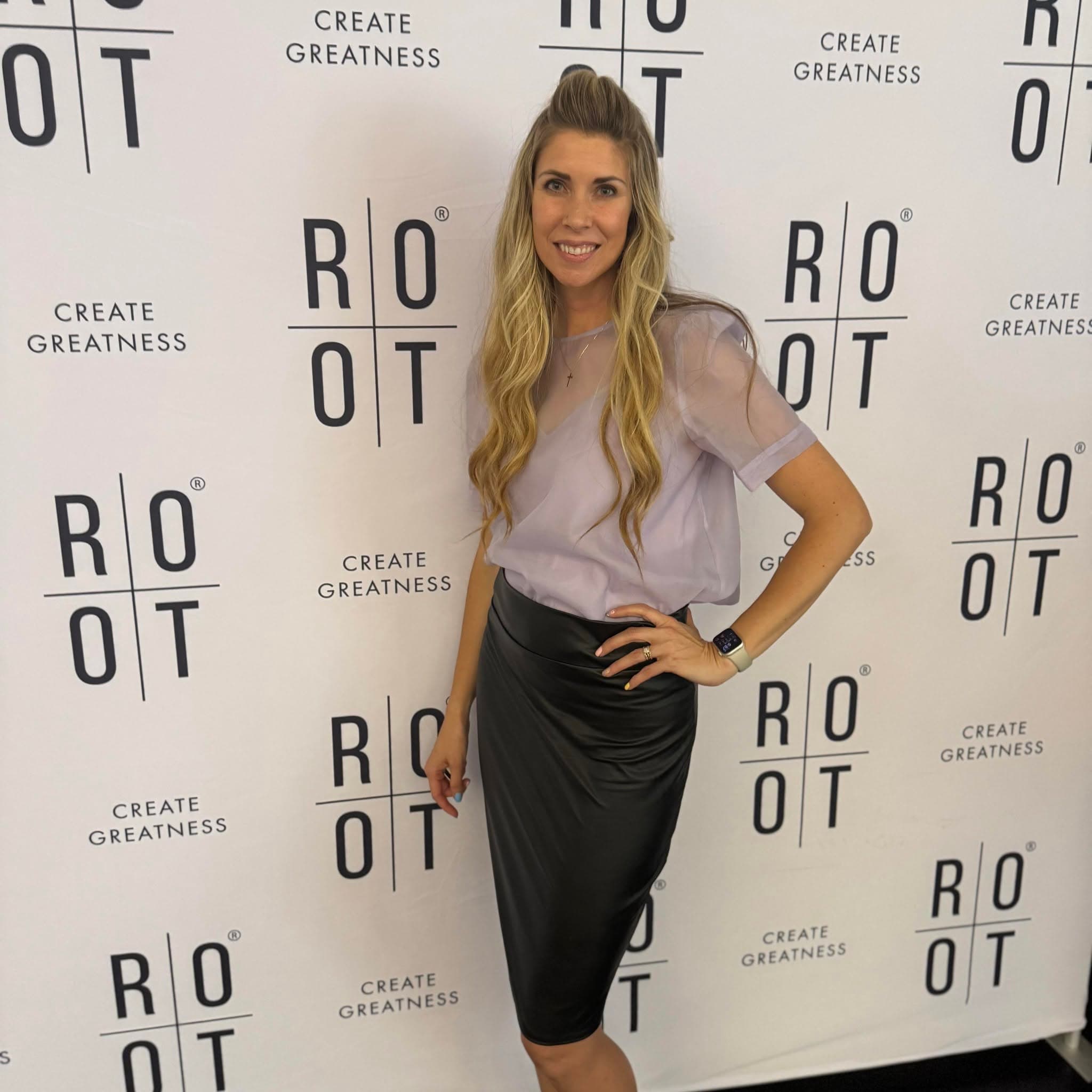 Smiling woman in lavender top and black skirt posing against a ROOT brand backdrop.