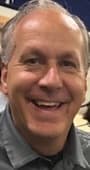 A smiling middle-aged man with short gray hair wearing a gray collared polo shirt.